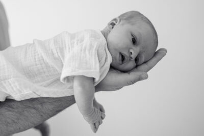 En noir et blanc, papa tenant son nouveau-né sur une paume ouverte, bébé en body de gaze blanche regardant l'objectif