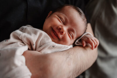 Nouveau-né souriant dans son sommeil couché sur l'avant-bras de son papa, body blanc à petites fleurs, ambiance douce