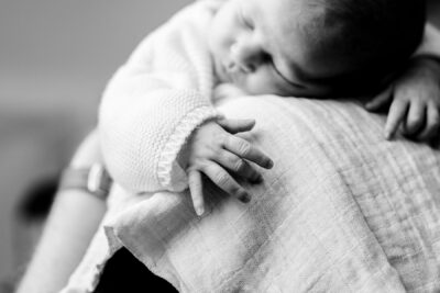 En noir et blanc, gros plan de la petite main d'un nouveau-né posée sur l'épaule de sa maman, bébé en pull tricoté blanc