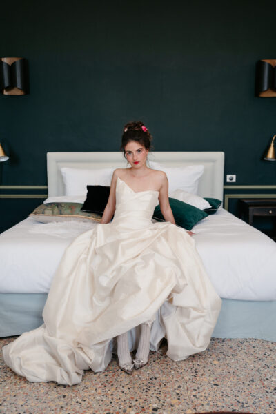 Mariée en robe bustier ivoire assise sur un lit dans une chambre aux murs vert foncé, roses roses dans les cheveux