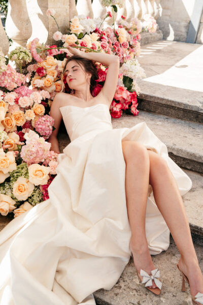 Mariée allongée sur des marches fleuries de roses pêche et rouges, robe bustier satin ivoire, escarpins à nœud strass