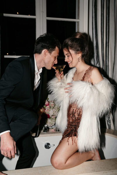 Couple de soirée partageant un martini, elle en mini-robe dorée paillettes et manteau en plumes blanches, sourires complices