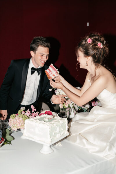 Mariée tendant une part de gâteau framboise à son mari en smoking, tous deux riant autour d'une table fleurie de roses roses