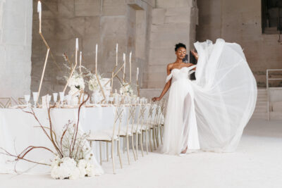 Mariée au maquillage doré faisant voler son voile près d'une longue table blanche dans un lieu de mariage en Île-de-France