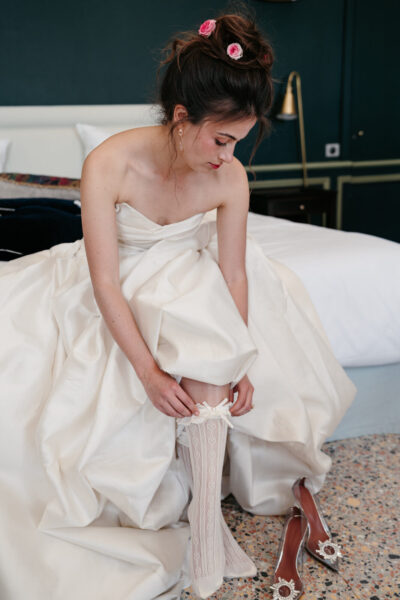 Mariée enfilant sa jarretière en dentelle assise sur un lit, bustier satin ivoire et roses roses dans le chignon