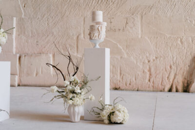 Gâteau de mariage à trois étages blanc et crème sur piédestal entouré de fleurs blanches et branches, mur en pierre de taille