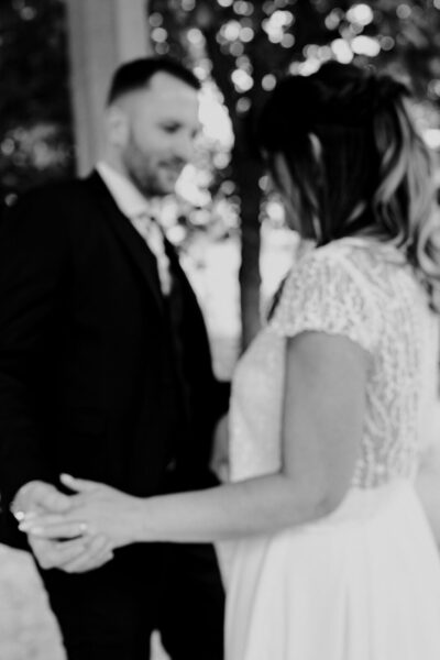 En noir et blanc, mariés face à face se tenant les mains sous un bokeh de lumières scintillantes, regard intime