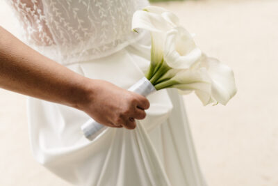 Gros plan d'une main de mariée serrant un bouquet de callas blancs noué d'un ruban satiné, robe dentelle en fond