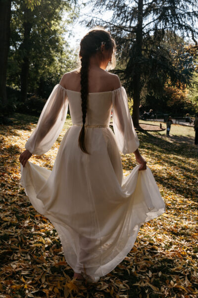 Mariée de dos faisant virevolter sa robe blanche à manches bouffantes sur un tapis de feuilles dorées dans un parc d'automne