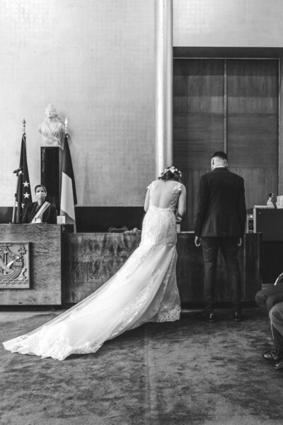 En noir et blanc, mariés de dos devant l'officier d'état civil en mairie, longue traîne de dentelle au sol
