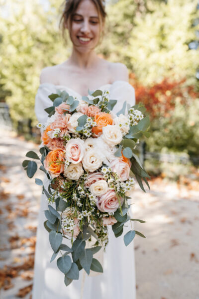 Mariée tenant un bouquet cascade de roses orange, pêche et blanches avec des feuilles d'eucalyptus