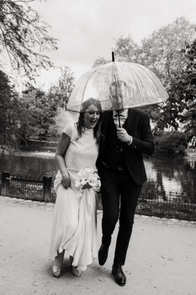 Mariés riant sous un parapluie transparent en bord d'étang dans un parc, ambiance festive sous la pluie
