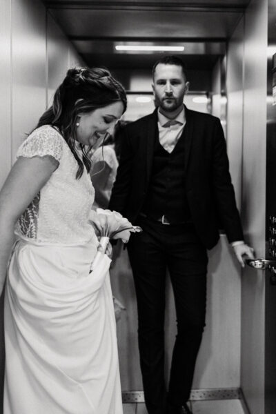 Mariée riant aux éclats dans un ascenseur, marié derrière elle l'observant avec tendresse