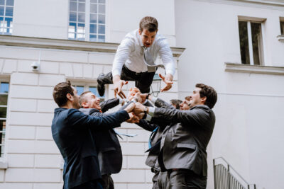Amis en costume propulsant joyeusement le marié en l'air devant un bâtiment haussmannien parisien