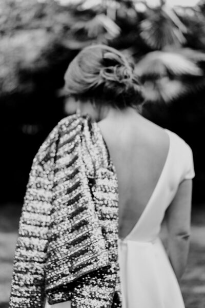 Mariée de dos en robe dos nu portant une veste à paillettes, chignon orné d'une barrette fleurie