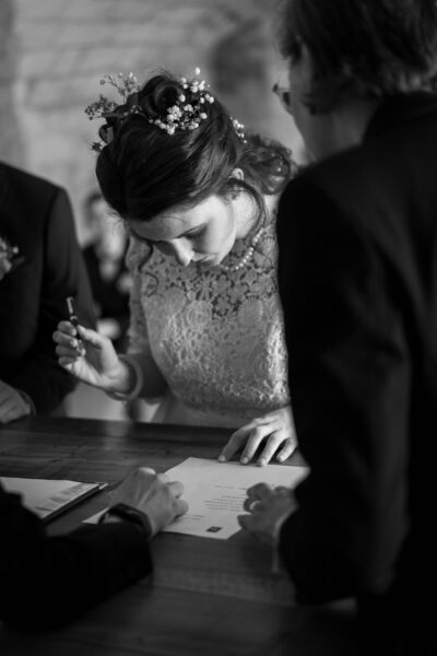 Mariée en robe de dentelle signant le registre de mariage, fleurs dans les cheveux, ambiance en N&B