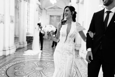 Mariée rayonnante traversant une galerie dorée en robe brodée, bouquet de pivoines à la main