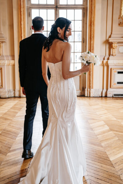 Mariée de dos en robe corset ivoire brodée tenant des pivoines blanches dans un salon haussmannien doré