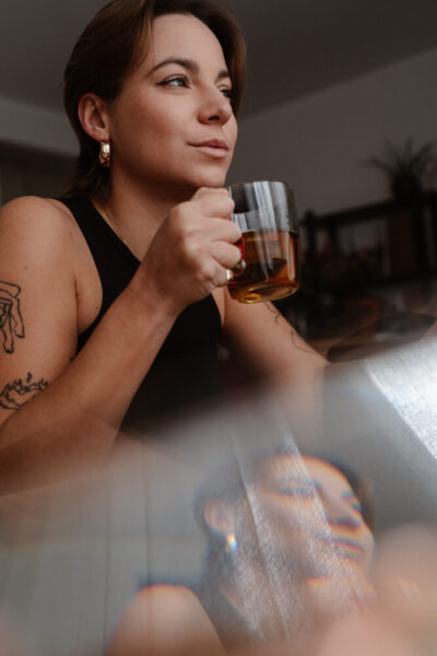 Femme tatouée en nuisette noire tenant un verre de thé ambré, regard mélancolique en lumière tamisée