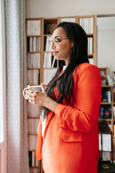 Femme en tailleur orange tenant une tasse de café debout face à une fenêtre lumineuse, regard pensif