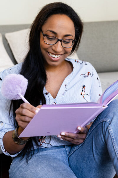 Femme aux lunettes riant en écrivant dans un carnet lilas avec un stylo pompon, cadre cosy en IDF