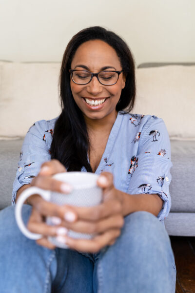 Femme aux lunettes souriant les yeux baissés vers une tasse de café entre ses mains, ambiance détendue