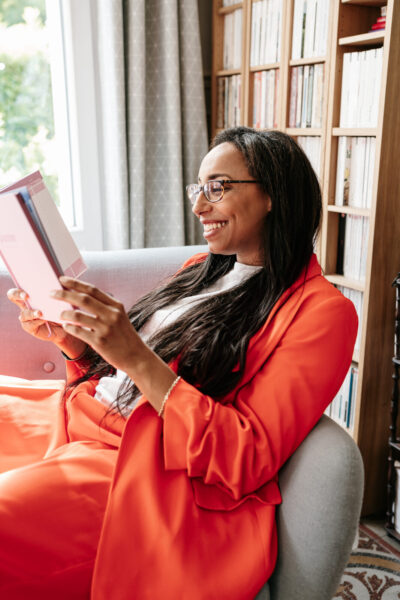 Femme en costume orange souriant en lisant un livre dans un fauteuil devant une grande bibliothèque