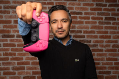 Homme en pull noir tendant une manette rose fluo vers l'objectif sur fond de briques industriel