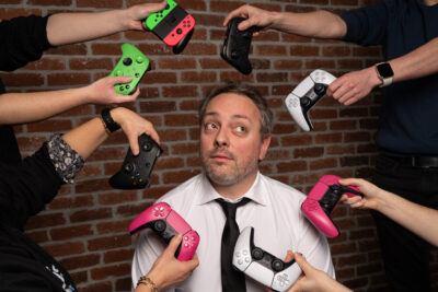 Homme en chemise et cravate encerclé de manettes de jeux vidéo colorées dans une mise en scène décalée