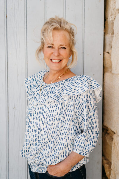 Femme blonde souriante en chemisier bleu à froufrous adossée à un mur en planches blanches