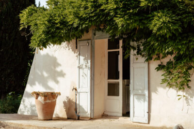 Façade blanche d'un mas provençal avec volets ouverts, vigne grimpante et bac en terre cuite au soleil