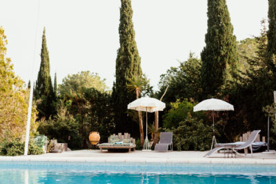 Piscine extérieure avec transats et parasols à franges blanches entourée de cyprès sous un ciel clair